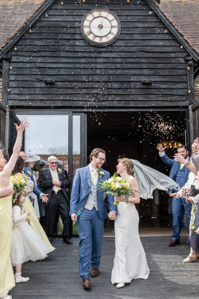 A bride and groom smiling and walking out of Lains Barn with guests throwing confetti, celebrating their wedding.