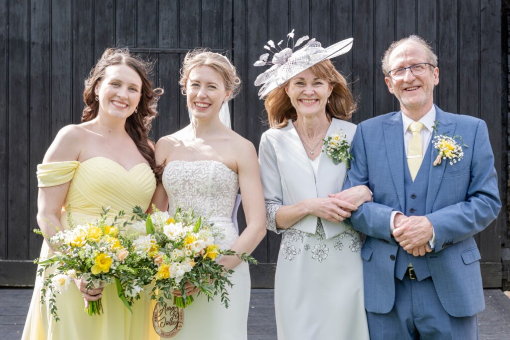 Four people at a wedding, with a bride and bridesmaid in the center holding bouquets, flanked by an older couple, with the woman wearing a hat. They are standing in front of a dark wooden background, all smiling.