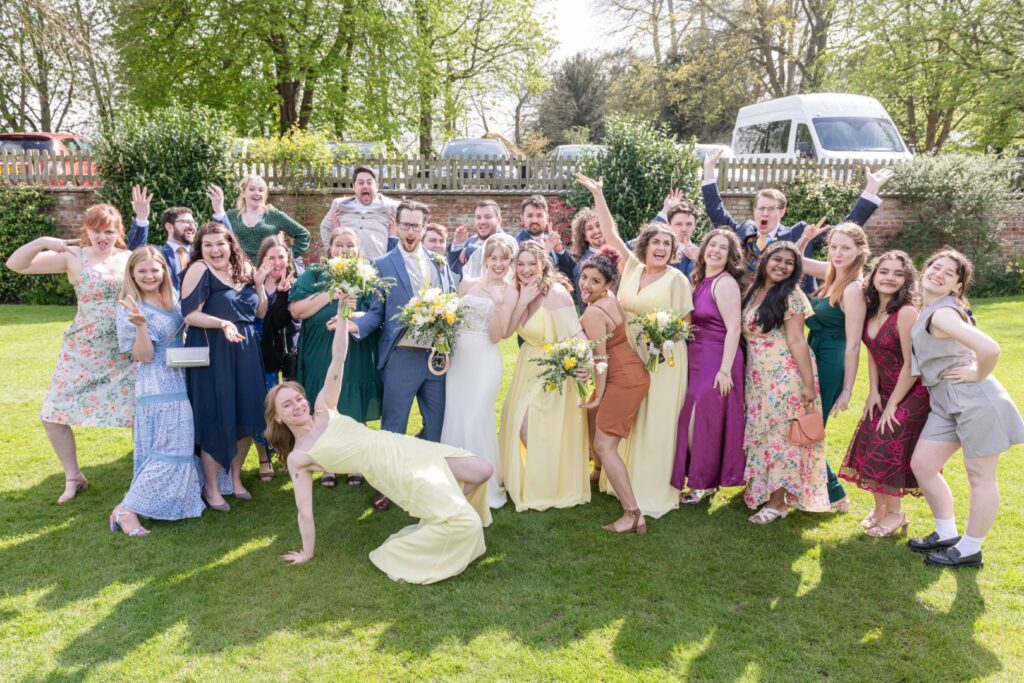 A vibrant group of people in formal wear, posing outdoors with a bride and groom at a wedding, some waving and smiling at the camera with a white van and greenery in the background.