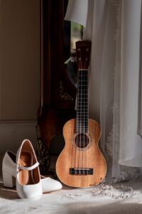 A ukulele leaning against a vintage dresser beside white heeled bridal shoes, wedding dress, veil and a delicate hair accessory on a soft surface with natural lighting.