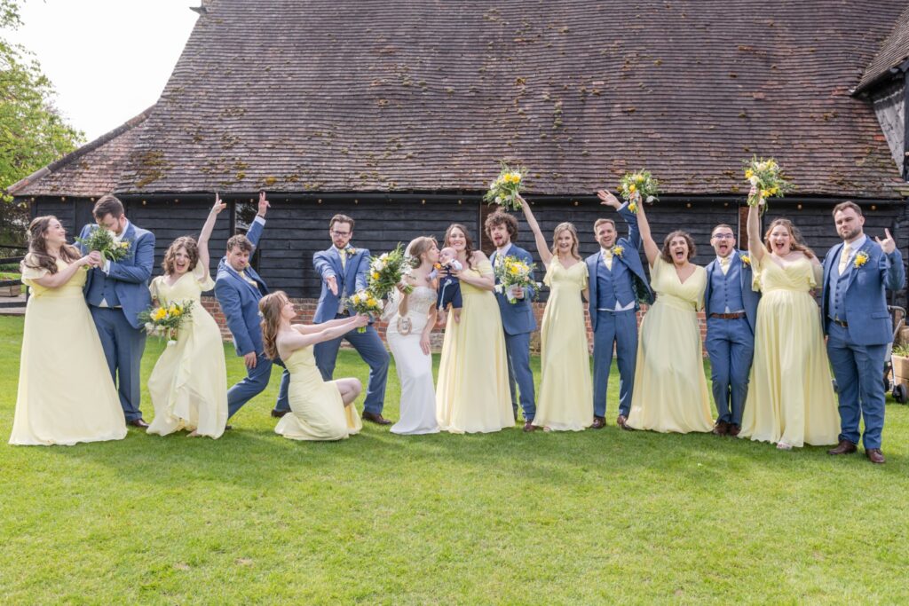 Group of happy people in formal attire celebrating at an outdoor wedding, with some of them holding bouquets and posing playfully on a lawn in front of a rustic building.