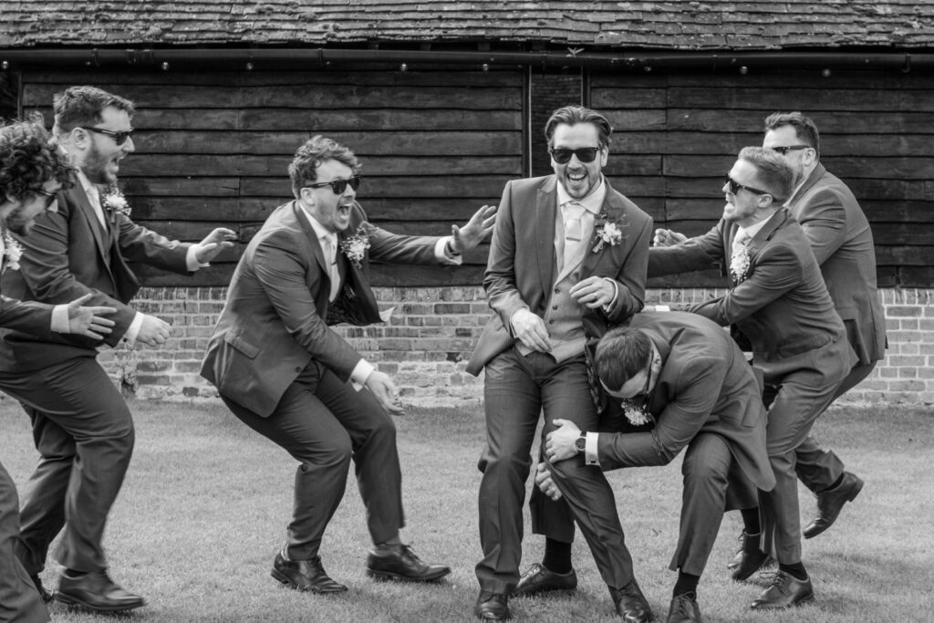 A black and white photo showing a group of groomsmen playfully fighting and laughing outdoors.