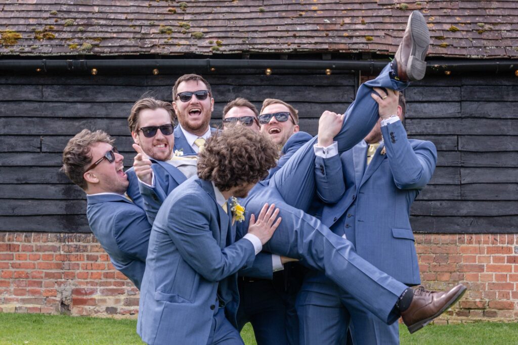 A group of groomsmen in blue suits laughing and playfully lifting the groom upside down at a wedding, with a rustic brick wall and roof in the background.