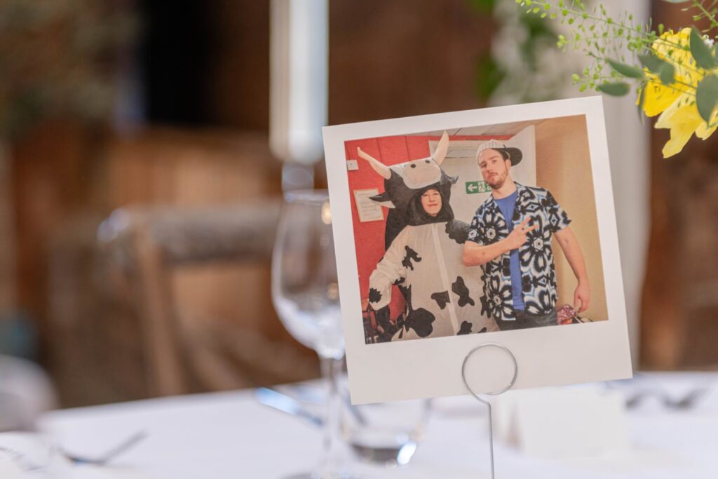 Photograph of two people in playful costumes displayed on a table with a blurred background of glasses and flowers.