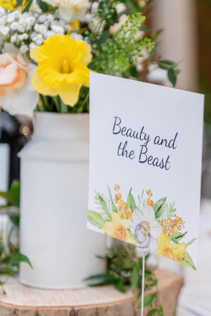 Close-up of a 'Beauty and the Beast' themed table sign with floral illustrations, displayed at an event, with blurred flowers and decorations in the background.