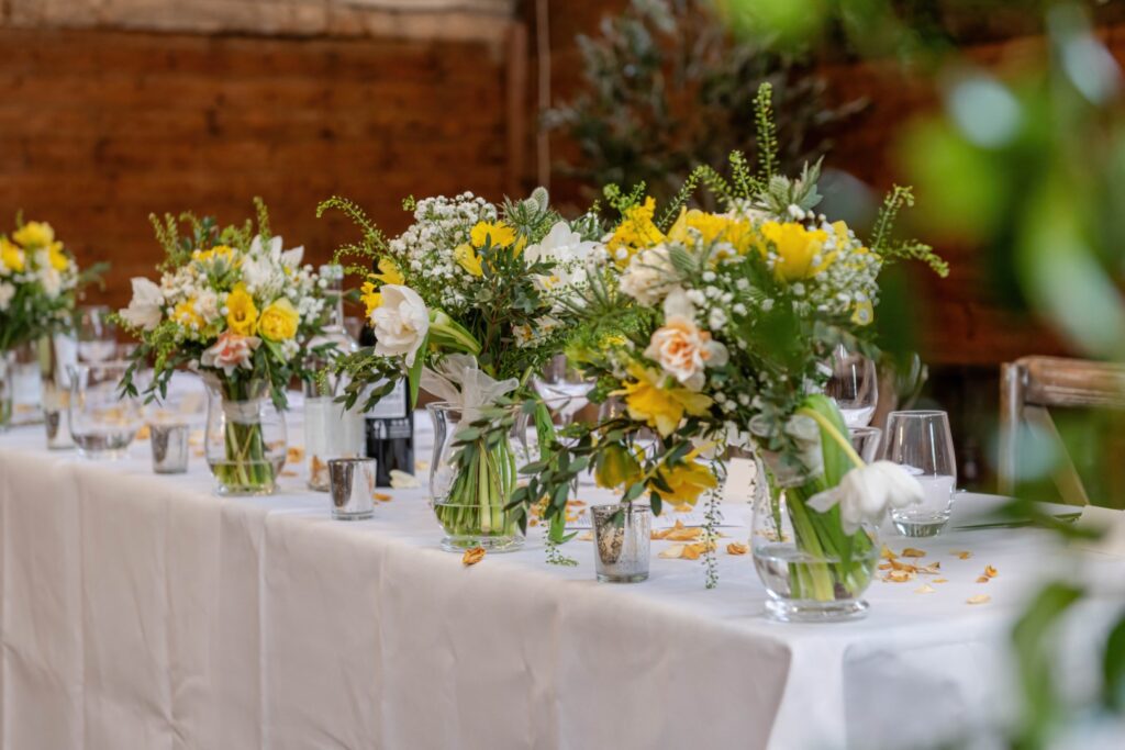 Several vases with fresh flowers arranged as centerpieces on a banquet table, with glasses and scattered petals.