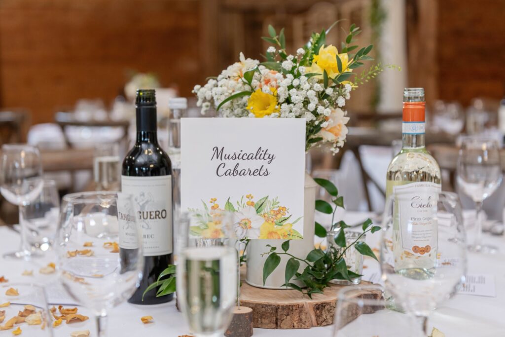 Close-up of an event table with floral centerpiece, wine bottles, and a place card that reads "Musicality Cabarets." Clear glassware and scattered rose petals complete the sophisticated decor.