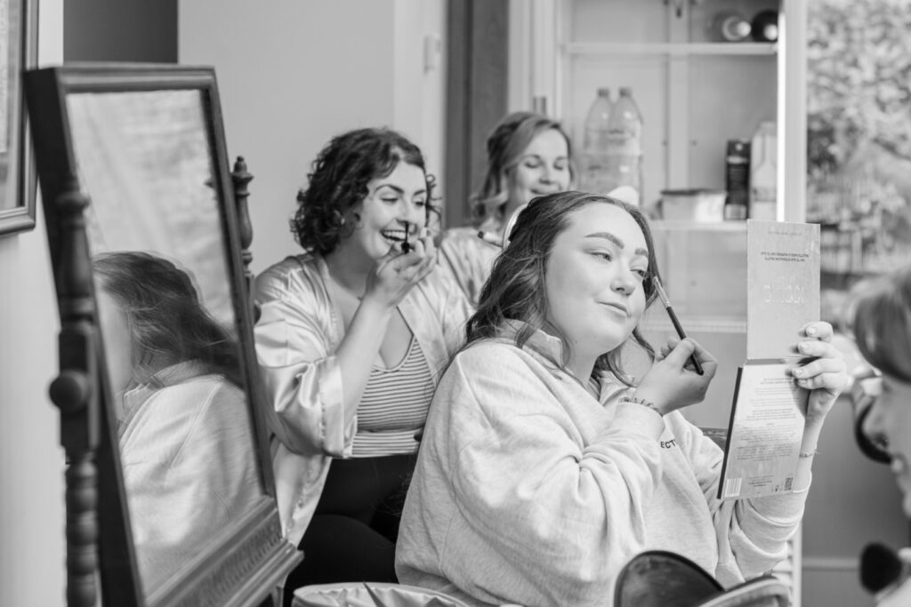 Black and white image of three laughing women in a room, two applying makeup while looking into handheld mirrors, capturing a candid moment of camaraderie.