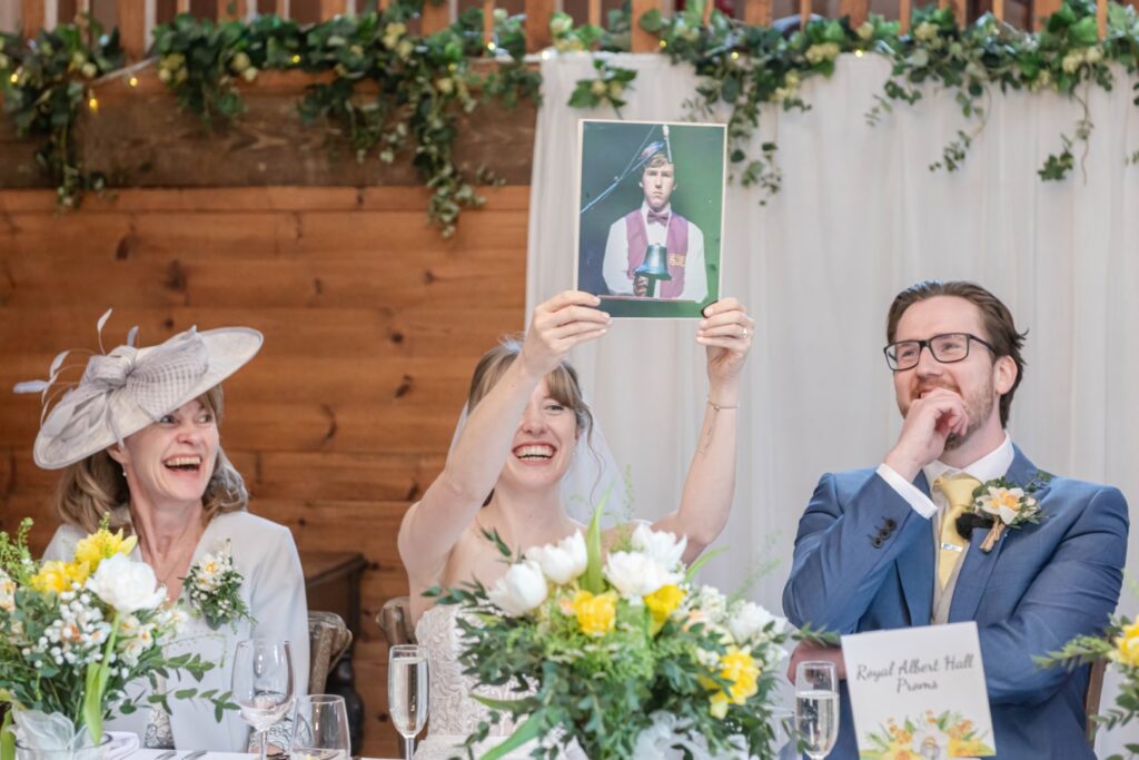 A bride holding up a framed photo smiling, next to a laughing woman in a hat and a smiling man in a blue suit at a wedding reception table decorated with flowers.