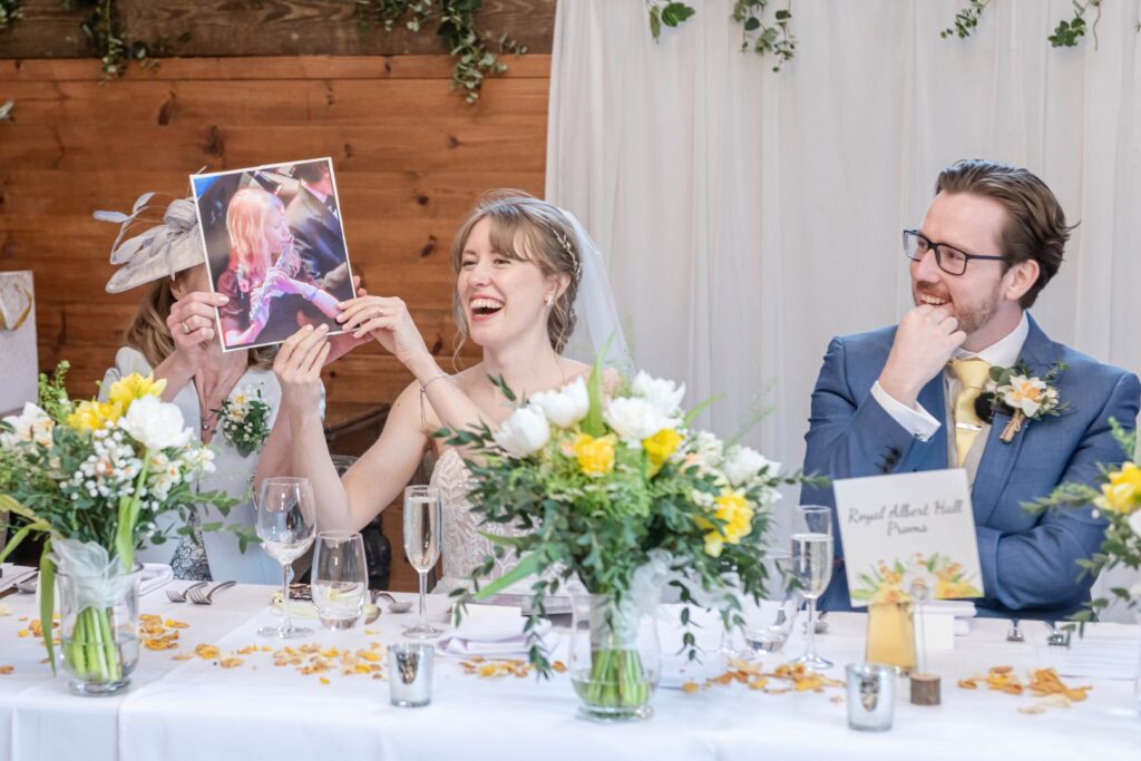 A bride laughing while holding up a photo covering her face, with a groom smiling at her, both sitting at a decorated wedding reception table.