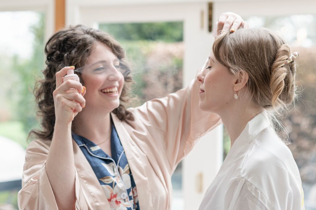 A smiling woman styling another woman's hair and applying hairspray in a well-lit room.