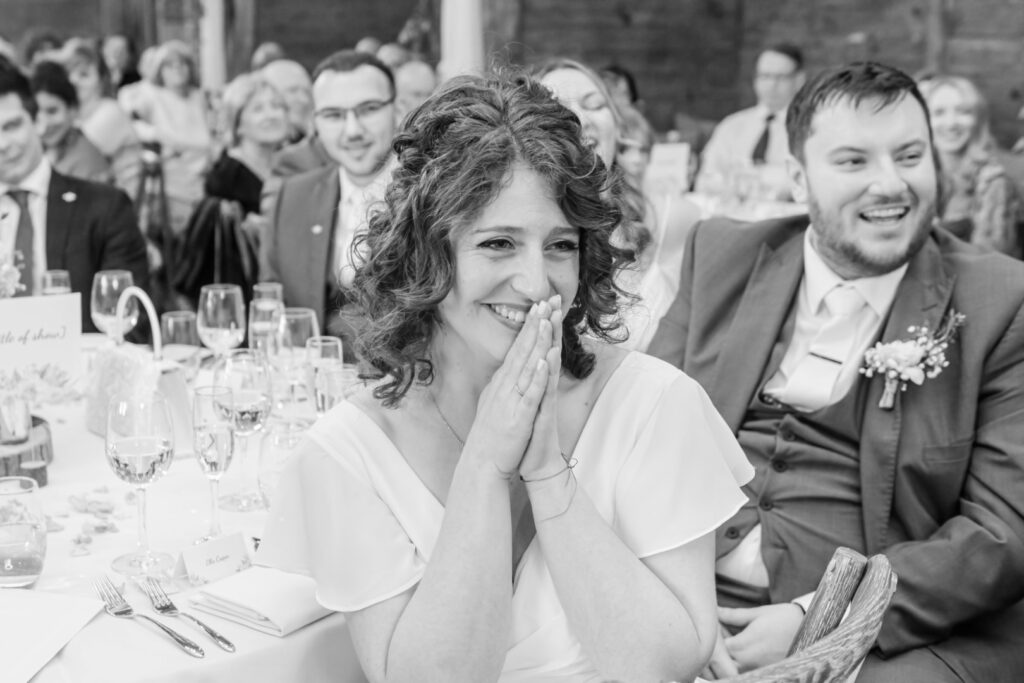 Black and white image of a smiling woman with hands pressed together, surrounded by happy guests at a wedding reception.