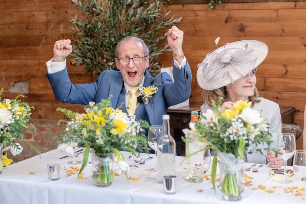 An elderly man in a blue suit excitedly raising his fists in victory while sitting at a wedding reception table with a smiling woman in a white outfit and a wide-brimmed hat beside him, surrounded by floral decorations and empty glasses.