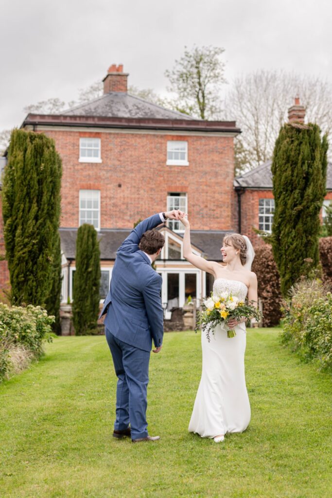 A bride and groom sharing a playful dance on a lawn with a brick house in the background.