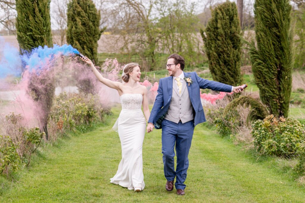 Bride and groom holding hands and smiling while walking with colorful smoke bombs in a garden setting.