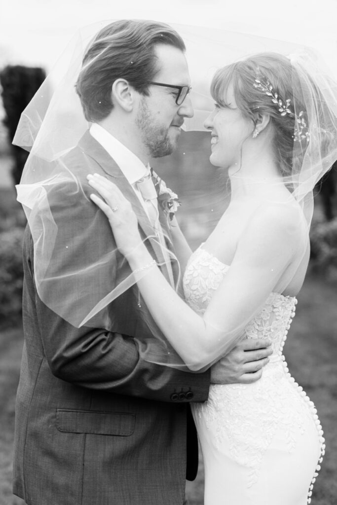 A black and white photograph of a bride and groom sharing a tender moment, embracing under the bride's veil.