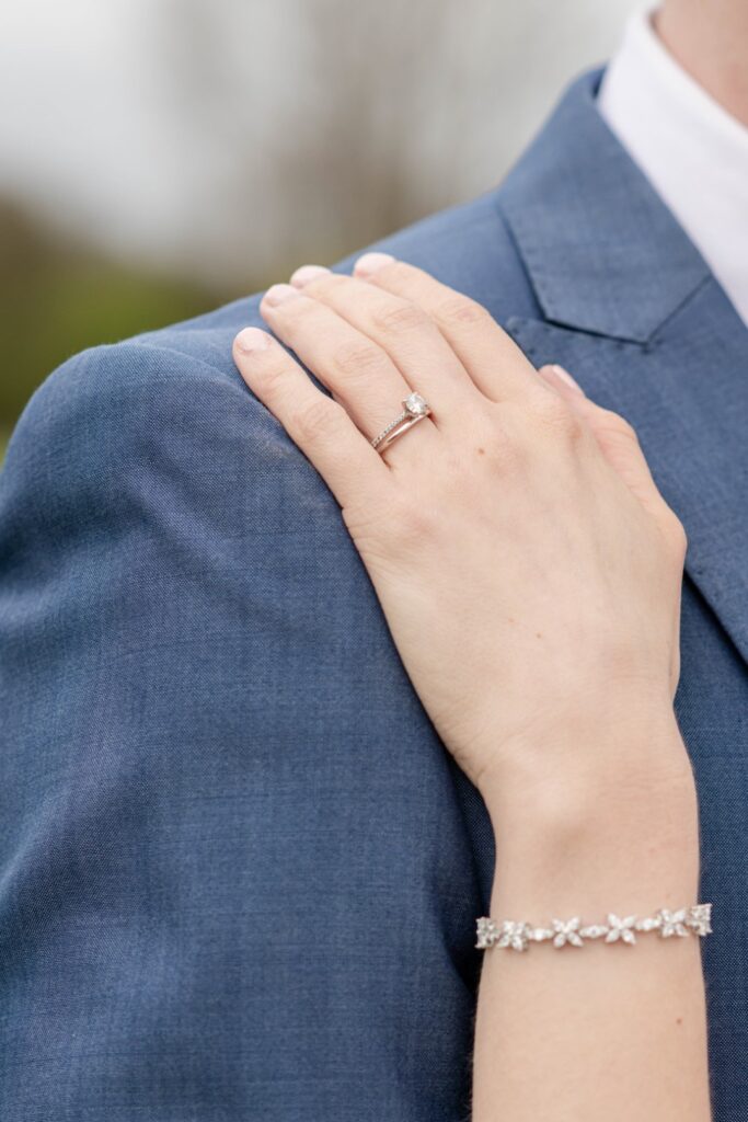 Close-up of a woman's hand resting on the shoulder of a person in a blue suit, featuring a prominent engagement ring and a floral bracelet.