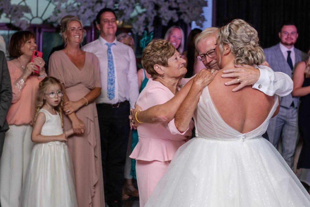 A bride in a white dress sharing a heartfelt dance with an older woman in pink, surrounded by smiling guests at a wedding reception.