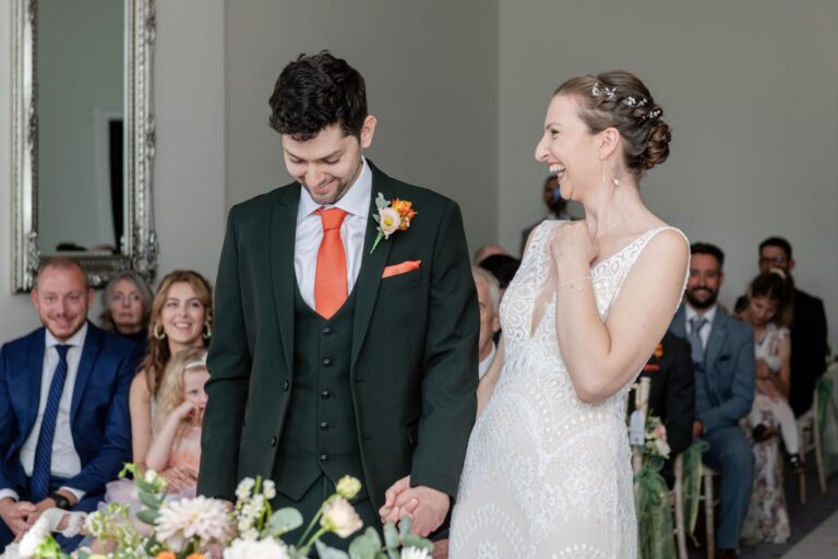 A bride and groom smiling at each other during their wedding ceremony with guests in the background.