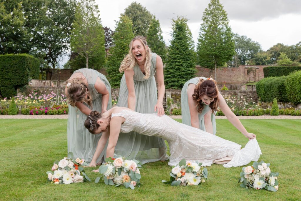 Bridesmaids' Laughter at Wedding:::Four women in formal wedding attire laughing and enjoying themselves in a garden setting, with the bride playfully leaning forward and bridesmaids holding her dress.