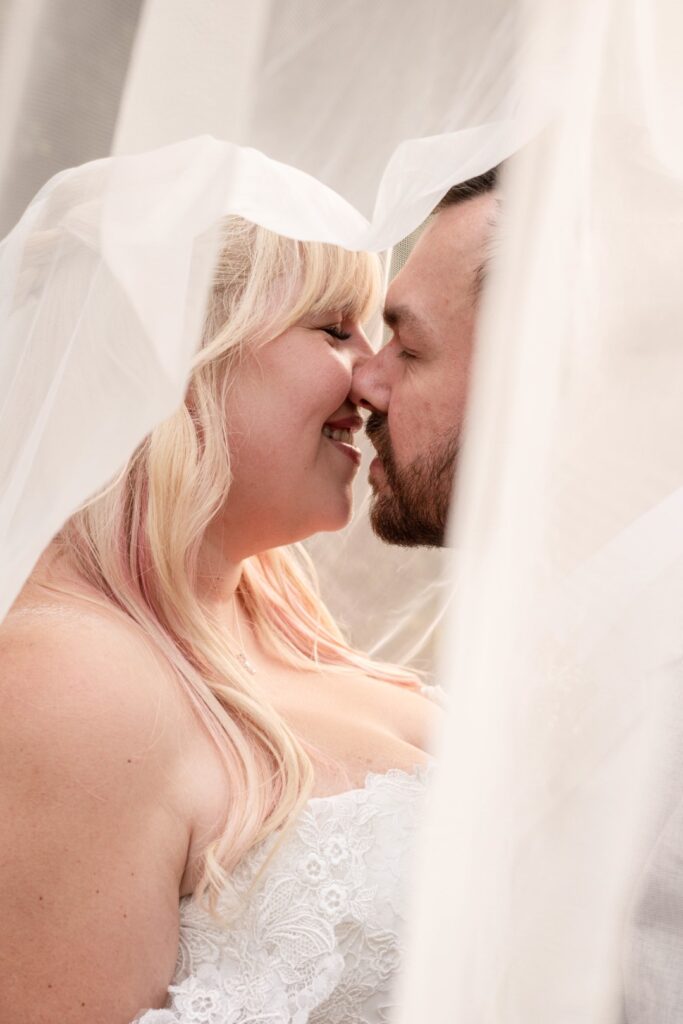 A bride and groom share a close moment, their faces partially covered by the bride's translucent wedding veil, with a tender expression of love and affection on their faces.