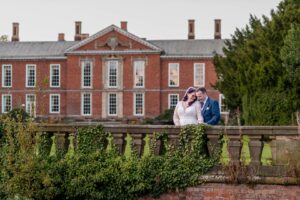 A bride and groom sharing an intimate moment by a stonewall balustrade with a grand historic house in the background.