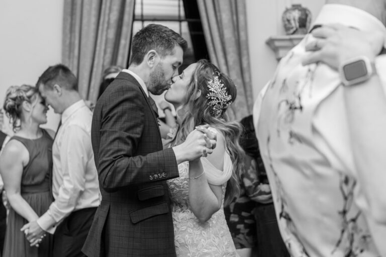 Black and white photo of a bride and groom sharing a kiss while dancing, surrounded by guests.