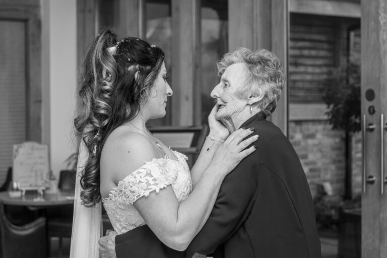 A black and white photo of a young bride in a lace dress embracing an elderly lady in a black outfit, both looking at each other with affectionate expressions.