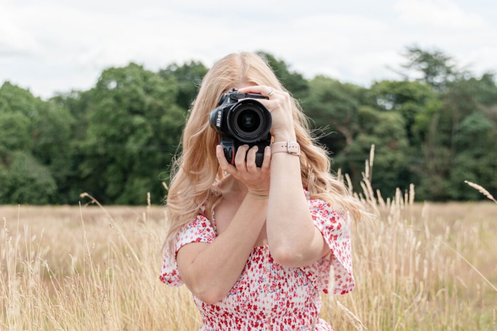 A woman with blonde hair taking a photo with a DSLR camera in a field of tall grasses.