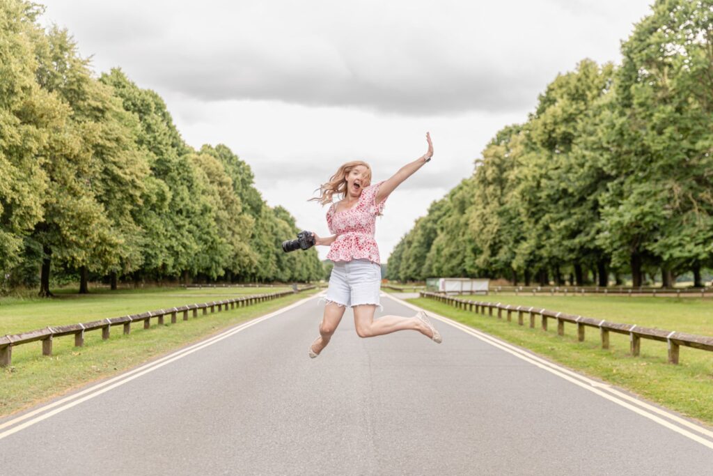 A woman joyfully jumping mid-air on a desolate tree-lined street, holding a camera and expressing happiness.