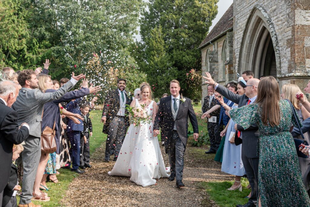 A joyful couple walks hand in hand, surrounded by guests showering them with confetti outside a charming stone church.