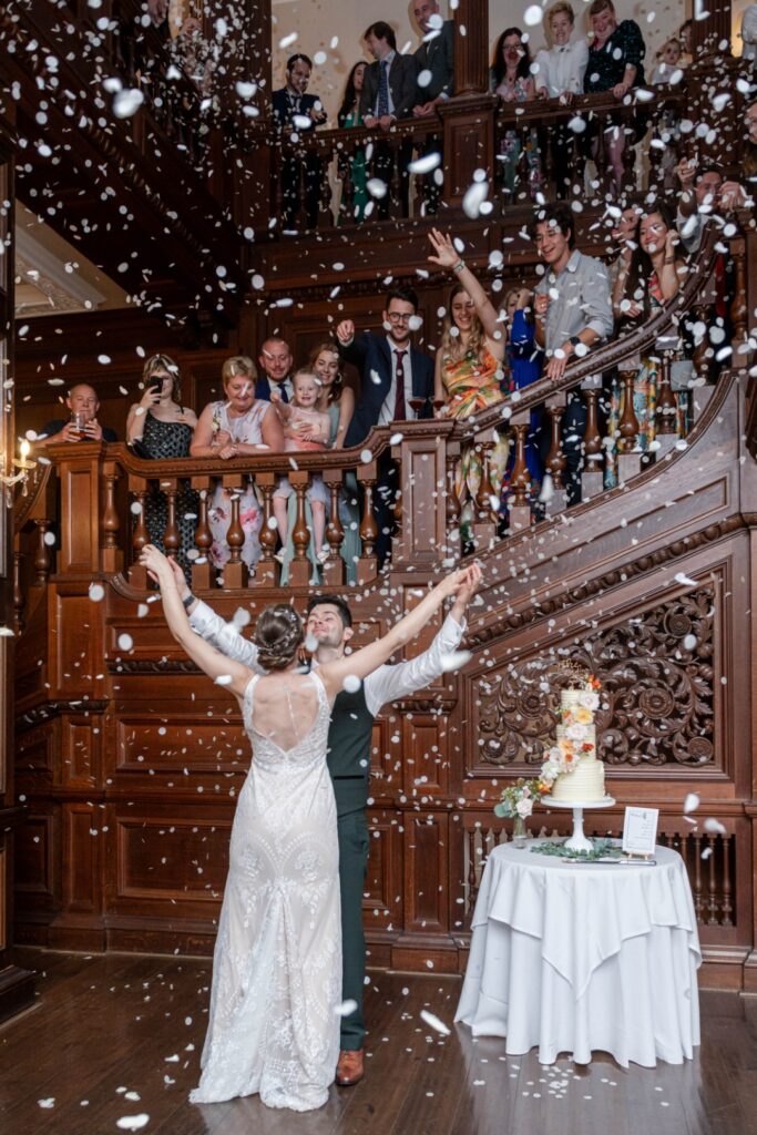 A joyful couple dances under falling confetti as guests smile and cheer from the balcony in a beautifully decorated venue.