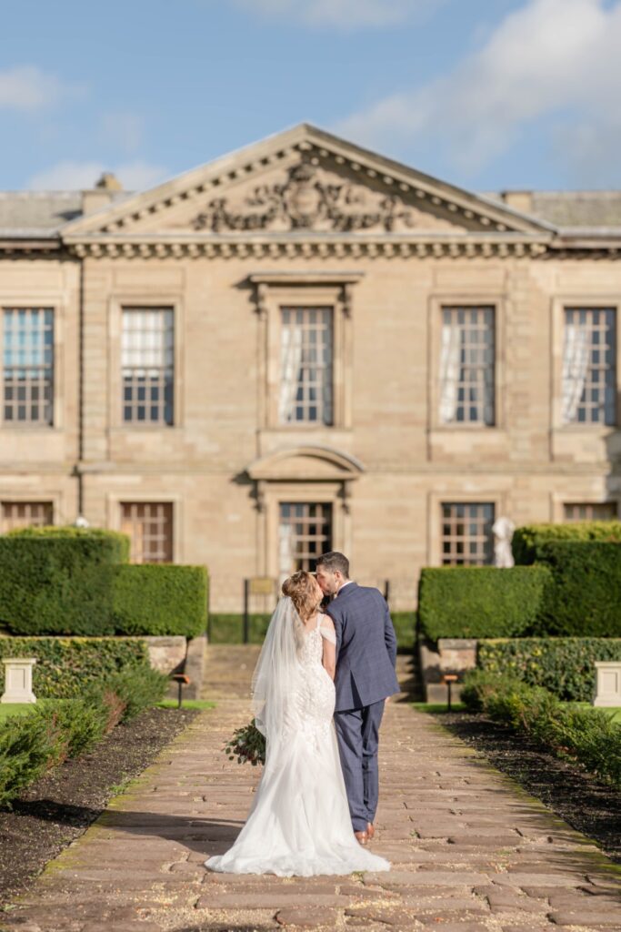 A couple in formal wedding attire shares a sweet kiss while standing in a beautifully landscaped garden in front of a historic stone building.