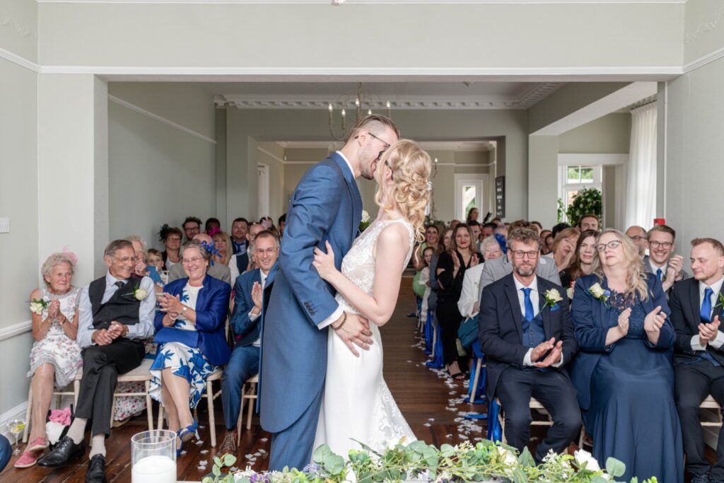 A couple shares a kiss during their wedding ceremony, surrounded by a cheering audience in a beautifully decorated venue.