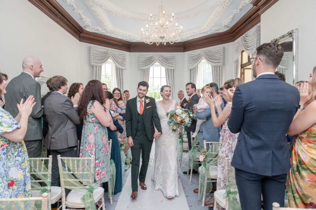 A bride and groom joyfully walk down the aisle hand-in-hand, surrounded by applauding guests in a beautifully decorated venue.