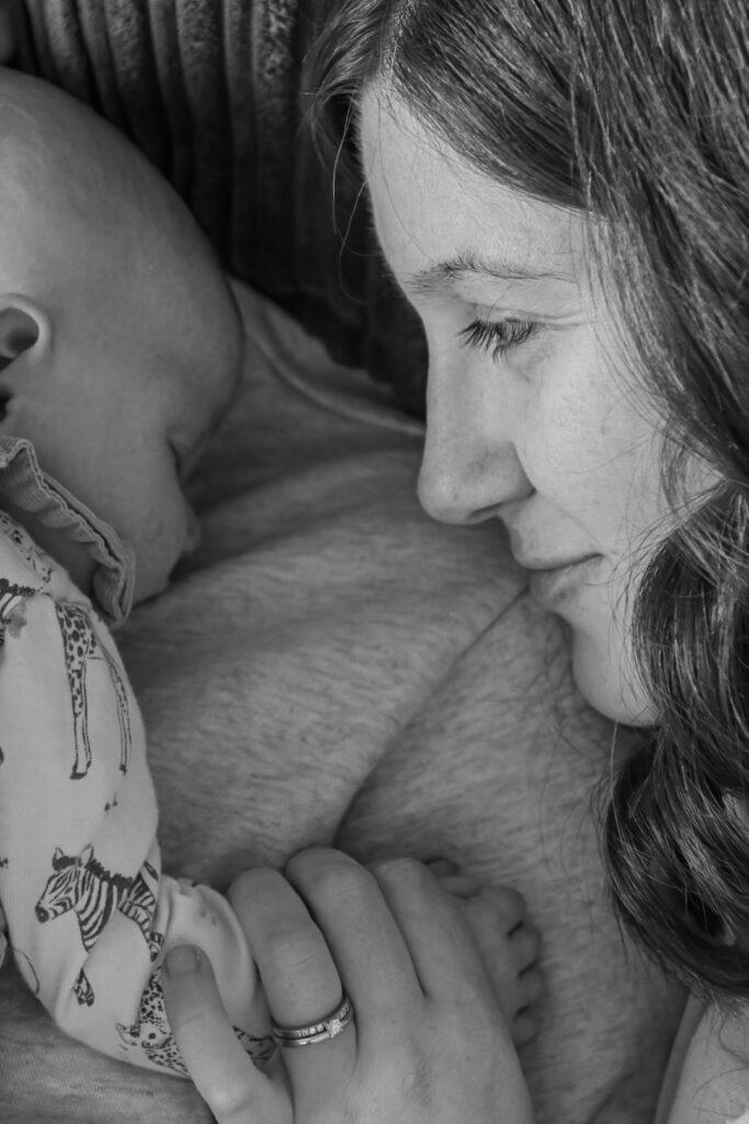 A close-up of a mother lovingly gazing at her sleeping baby, both resting against a textured background. The mother’s hand tenderly holds the baby's hand, showcasing a moment of warmth and connection.