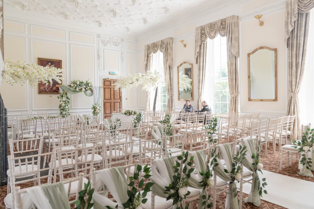 A beautifully arranged indoor wedding ceremony space featuring rows of chairs adorned with fabric and greenery, complemented by floral decorations and ornate architectural details.