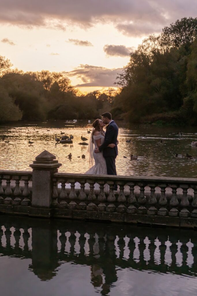 A couple shares a tender kiss by a serene lake during sunset, surrounded by lush trees and swimming ducks, reflecting love and tranquility.