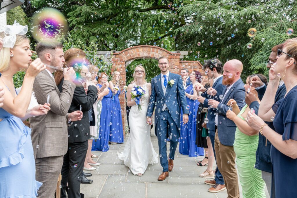 A joyful couple walks hand-in-hand as friends and family blow bubbles in celebration, surrounded by bridesmaids in blue dresses and a picturesque garden backdrop.