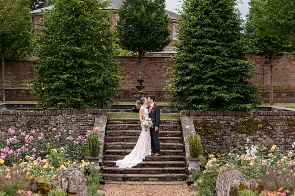 Couple sharing a kiss on stone steps surrounded by lush greenery and blooming roses in a picturesque garden.