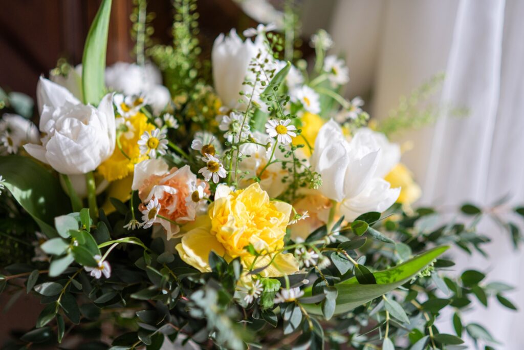 A close-up of a vibrant bouquet featuring white tulips, yellow roses, and delicate daisies, surrounded by greenery, illuminated by natural light.