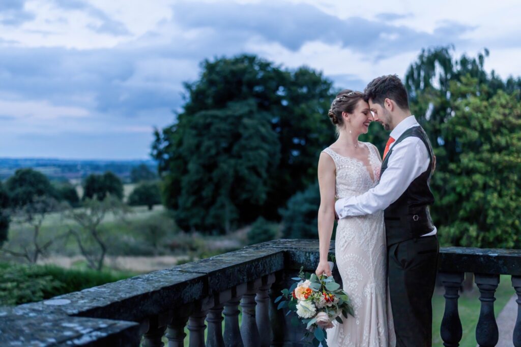 A couple shares a tender moment on a balcony, surrounded by lush greenery and a scenic landscape at twilight.