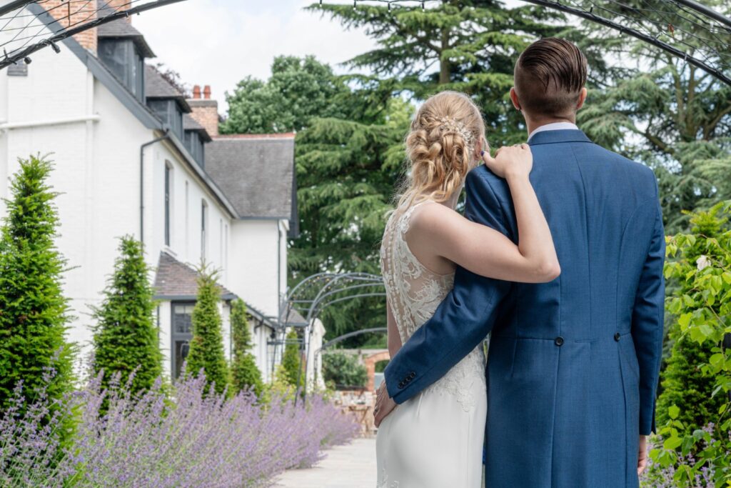 A couple dressed in elegant wedding attire stands together in a beautiful garden setting, surrounded by lavender flowers and greenery, with their backs to the camera.