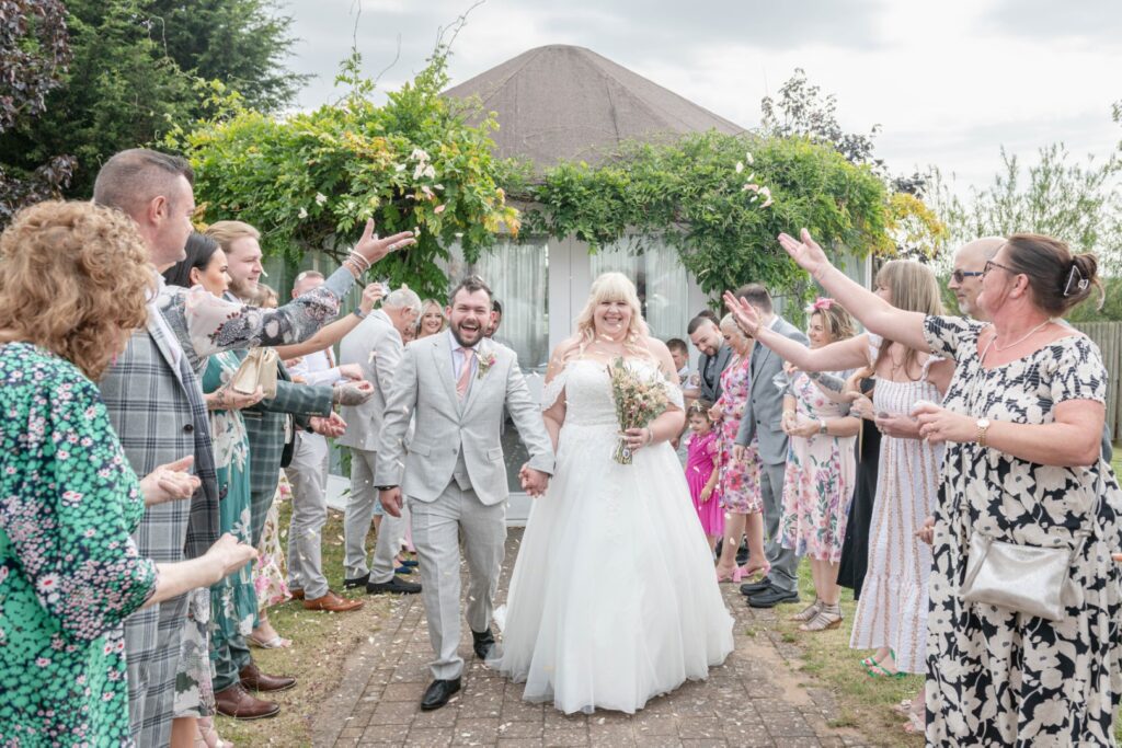 A happy couple walks hand in hand down a path surrounded by guests throwing confetti, celebrating their special day outdoors.