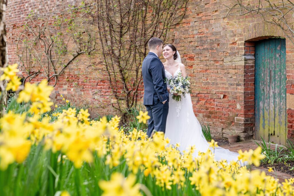 A couple shares a joyful moment in front of a rustic brick wall, surrounded by vibrant yellow daffodils and lush greenery.