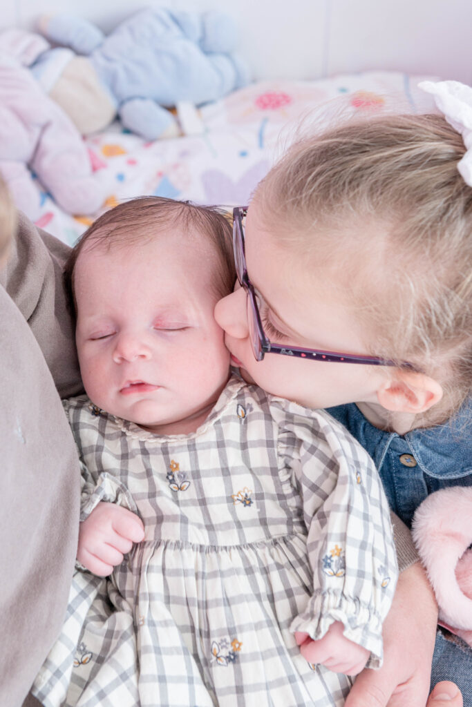 A young girl lovingly kisses her sleeping baby sister while being cradled in a cozy setting.