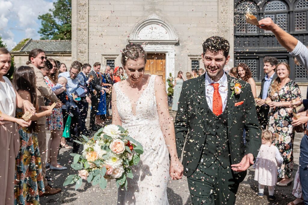 A couple walks hand in hand, showered with confetti, as their guests celebrate their union outside a beautiful venue.