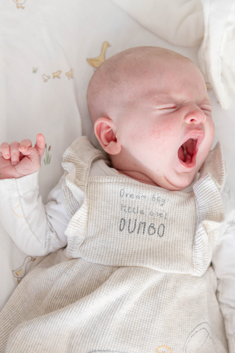 A baby yawning while lying on a soft bed, dressed in a beige outfit with the phrase "Dream big little one" embroidered on it.