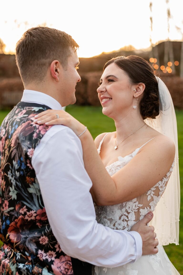 A joyful couple embraces during their wedding, with the bride in a floral lace gown and the groom in a colorful floral vest, smiling at each other against a picturesque sunset backdrop.