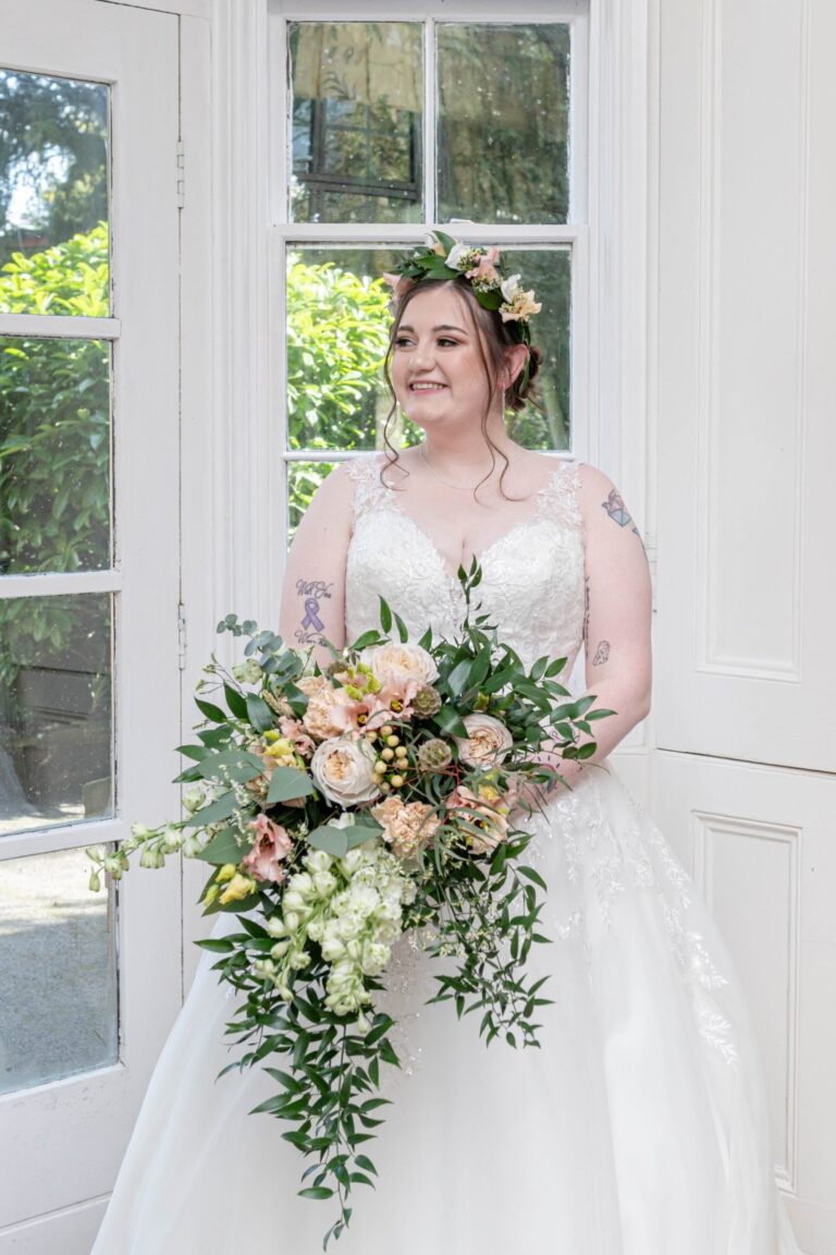 A smiling bride wearing a floral crown holds a lush bouquet, standing by a window with greenery outside.